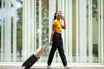 Cheerful woman talking on mobile phone while carrying luggage on city street