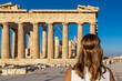 Rear view of tourist woman in white dress looking at Parthenon of Acropolis of Athens, Attica, Greece, Europe. Ruins of ancient temple, the birthplace of democracy. Girl wearing golden laurel crown