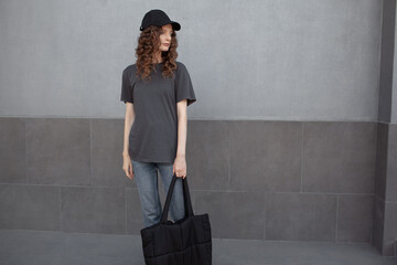 Wall Mural - A beautiful girl is wearing a grey t-shirt,black bag,  cloth cap and jeans posing against the background of a wall. fashion outfit for the city, minimalism urban style clothing