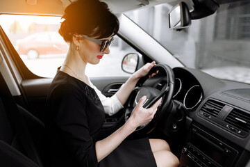 Attractive successful business woman sits inside of the luxury car and texting on her smartphone. Business owner woman in sunglasses driving a car solves work issues on the way to work.