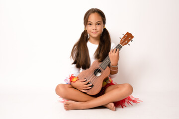 Wall Mural - Cute little girl in summer clothing playing ukulele sitting on the floor over white background. Happy vacation concept.