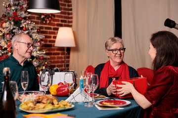 Family exchanging christmas gifts at festive table, celebrating winter holiday together, having dinner. Xmas celebration with parents, mother and father giving daughter present box