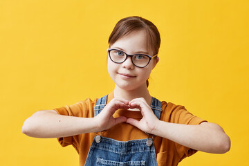 waist up portrait of teen girl with down syndrome looking at camera and showing heart sign while sta