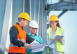 © Olga - young men and a woman are standing in orange and white helmets and signal vests with documents against the background of the metal structure of the bridge. The concept of railway and road engineers.