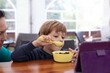 © Татьяна Волкова - Little boy sit on chair at table and watch cartoon on tablet computer with mother. Kid of kindergarten age eat chocolate balls with milk from children tableware. Ready breakfast, gadget addiction