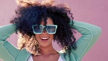 Black woman with sunglasses smile, play with hair and laugh against pink backdrop in sunshine. Model girl with curly afro hair, happy with blue fashion glasses against color wall or background