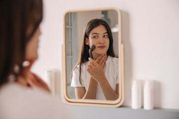 Young caucasian woman wears white t-shirt uses ball roller in front of mirror in her bedroom. Brunette hair female massages face with Y-shaped massager at home. Concept morning routine, facial massage