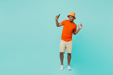 Wall Mural - Full size young man of African American ethnicity wear orange t-shirt hat hold in hand use mobile cell phone dance have fun isolated on plain pastel light blue cyan background People lifestyle concept