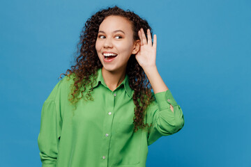 Wall Mural - Young curious nosy woman of African American ethnicity 20s she wear green shirt try to hear you overhear listening intently isolated on plain blue background studio portrait. People lifestyle concept.