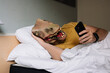 © karrastock - Man with lizard mask using the smartphone in a hotel bed.