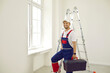 © Studio Romantic - Portrait of male worker inside house. Happy man in work uniform overalls standing in new empty room with white walls, holding toolbox, looking at camera and smiling. Repairs and renovations concept