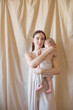 Young mother with long dark hair holding a cute little daughter in her arms in a nude fabric on a nude textile background. happy motherhood