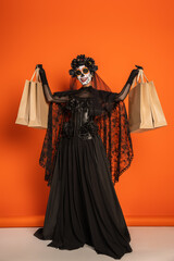 full length of cheerful woman in black witch dress and halloween makeup showing shopping bags on orange background.