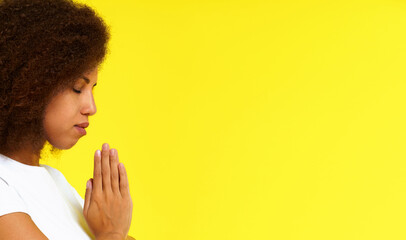 Young african american woman in prayer position wearing white t-shirt isolated on yellow background. African girl praying God studio shot. Spiritual woman put hands in prayer