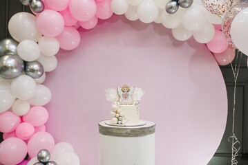 Birthday Cake on a background pink, white and silver balloons. Baby shower party. Trendy Cake with a figure angel for a girl. Copy space. Celebration baptism concept. Photo wall and arch with decor.