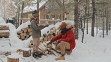 Young Adult Black Father In Winterwear Sitting On Squats At Woodpile Covered With Snow, Passing Firewood To His Son