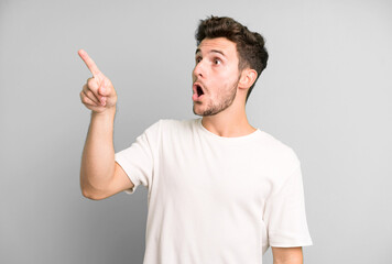 Wall Mural - young handsome man feeling shocked and surprised, pointing and looking upwards in awe with amazed, open-mouthed look