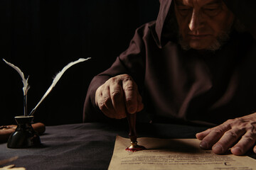 Poster - medieval monk in dark robe with hood stamping parchment with wax seal isolated on black.