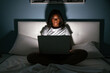 © David Prado/Stocksy - Smiling african woman using laptop in bed at night