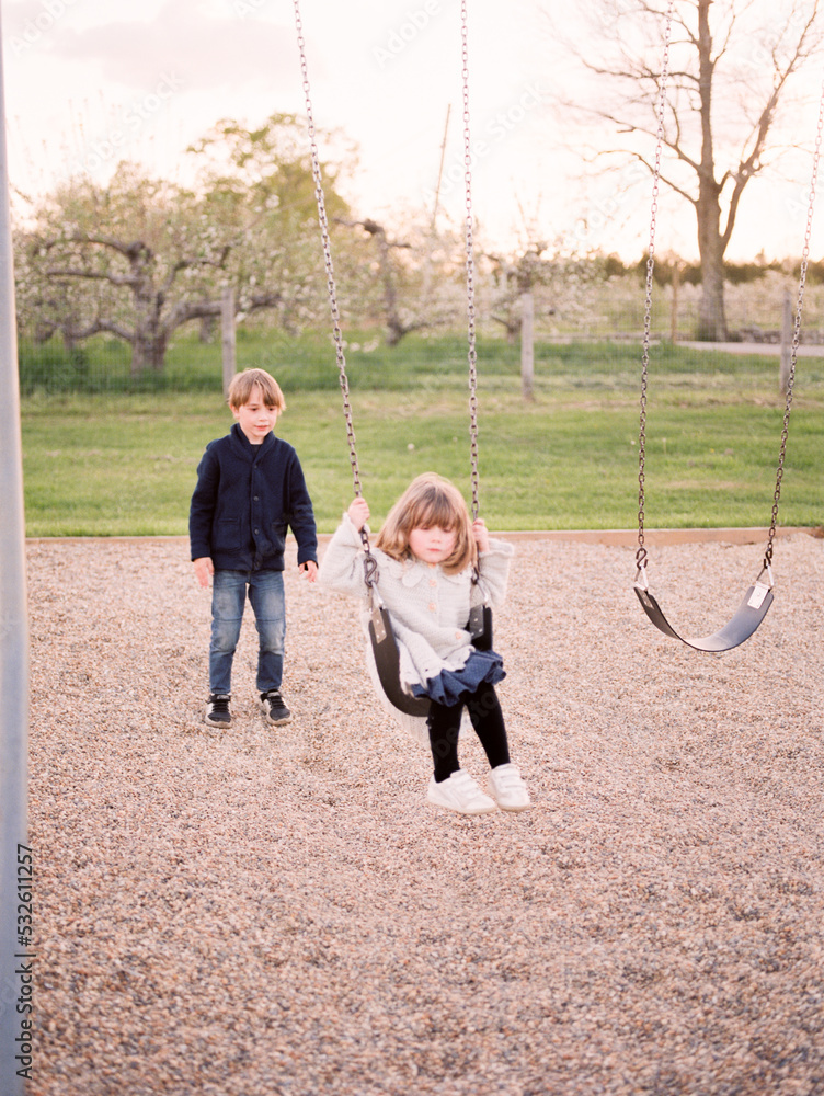 Little Boy Pushing His Little Sister On A Swing At A Playground Stock