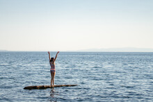 Girl Stands On Driftwood Log In Ocean Arms Raised In Victory
