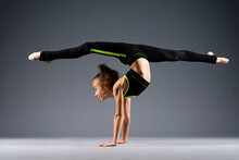 Flexible Little Girl Doing Acrobatic Stunts While Standing On Her Hands. The Gymnast Is Dressed In A Black Tracksuit For Stretching. Isolated On A Gray Background.