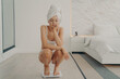 © VK Studio - Unhappy young caucasian female squatting on tiptoes over electronic scales in bedroom with sad expression
