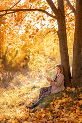 A young woman in a warm coat is sitting on autumn golden leaves and reading a book. A girl is resting in an autumn park