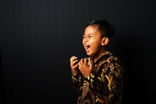An Asian Kid Wearing Batik Is Screaming Isolated On Black Background.