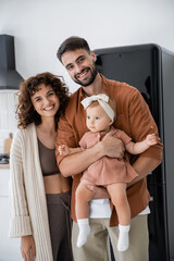 happy woman standing near bearded husband holding in arms baby daughter.