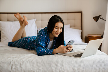 Wall Mural - Asian girl using laptop and mobile phone while lying in bed at home