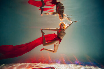 Art work. A slender, tanned girl with an athletic figure and blond hair, with red material and light underwear, in a ballet pose underwater in the pool. Aesthetic image for your design or decoration.