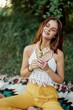 © SHOTPRIME STUDIO - Young hippie woman sitting in nature on a plaid with a flower in her hands and looking at the camera smiling sincerely in eco-clothing in autumn in the sunset