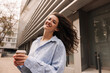 © Look! - Cute young caucasian girl holding coffee, waving her hair standing outdoors. Brunette in good mood wears blue shirt. Lifestyle concept