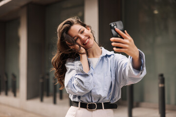 Wall Mural - Cute caucasian young girl communicates by video via smartphone standing near business center. Brunette with wavy hair wears blue shirt to street. Cell phone usage concept