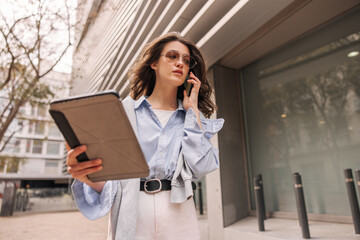 Wall Mural - Business pretty young caucasian woman uses modern technologies standing near office. Brunette wears classic clothes and sunglasses. Internet concept outside.