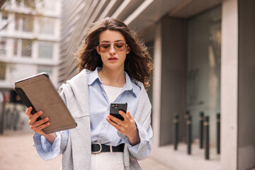 Wall Mural - Business young caucasian woman uses two wireless devices outdoors. Brunette with flying hair wears sunglasses, shirt, sweatshirt. Management concept