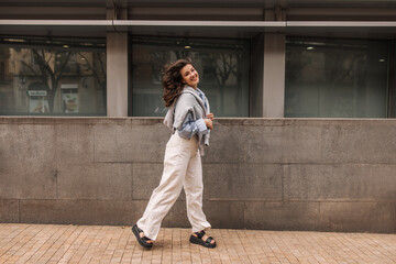 Wall Mural - Full length positive young caucasian girl in shirt and trousers on street near office. Brunette with wavy flying hair. Concept of journalism, learning.