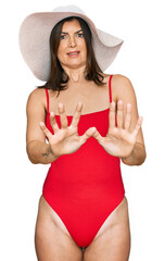 Wall Mural - Beautiful brunette woman wearing swimsuit and summer hat afraid and terrified with fear expression stop gesture with hands, shouting in shock. panic concept.