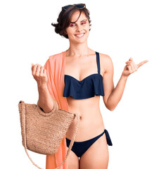 Poster - Beautiful young woman with short hair wearing bikini and hat holding summer wicker handbag smiling happy pointing with hand and finger to the side