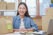 Small business entrepreneur SME, smile asian young woman, girl owner sitting confident on table before prepare order sent to customer packing box to delivery, working online at home, ecommerce.