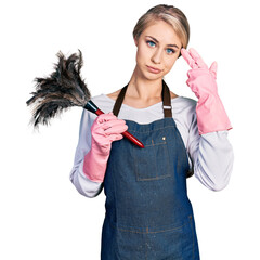 Wall Mural - Beautiful young blonde woman wearing apron holding cleaning duster shooting and killing oneself pointing hand and fingers to head like gun, suicide gesture.