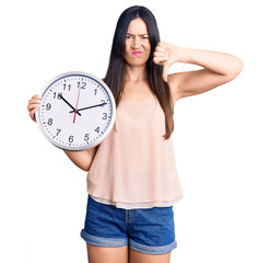 Sticker - Young beautiful caucasian woman holding big clock with angry face, negative sign showing dislike with thumbs down, rejection concept