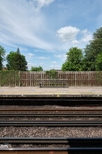 Train Station Platform With A Wooden Bench And Fence. Trees And Blue Sky With White Clouds In The Background.