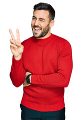 Wall Mural - Young hispanic man wearing casual clothes smiling with happy face winking at the camera doing victory sign with fingers. number two.