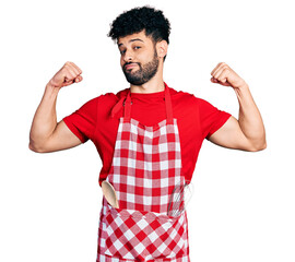 Poster - Young arab man with beard wearing cook apron showing arms muscles smiling proud. fitness concept.