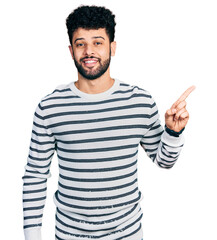 Sticker - Young arab man with beard wearing casual striped sweater with a big smile on face, pointing with hand finger to the side looking at the camera.