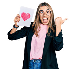 Canvas Print - Hispanic young woman holding heart draw pointing thumb up to the side smiling happy with open mouth