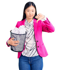 Sticker - Young beautiful chinese woman holding paper bin full of crumpled papers with angry face, negative sign showing dislike with thumbs down, rejection concept