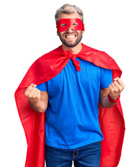 Canvas Print - Young blond man wearing super hero custome very happy and excited doing winner gesture with arms raised, smiling and screaming for success. celebration concept.